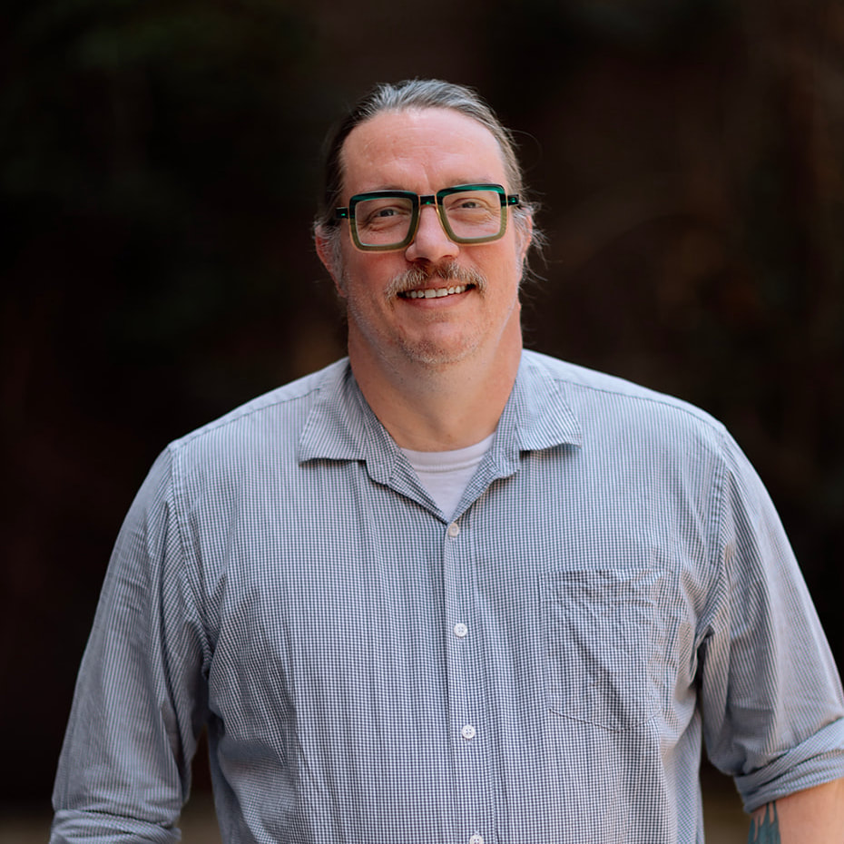 Our Team 14 Smiling man with pulled-back hair, mustache, and thick green glasses in a striped shirt.