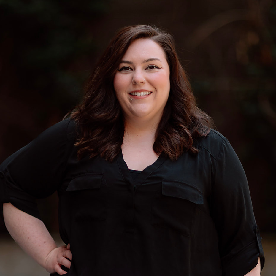 Our Team 9 Smiling woman with dark wavy hair, labret piercing, black shirt, and hand on hip.