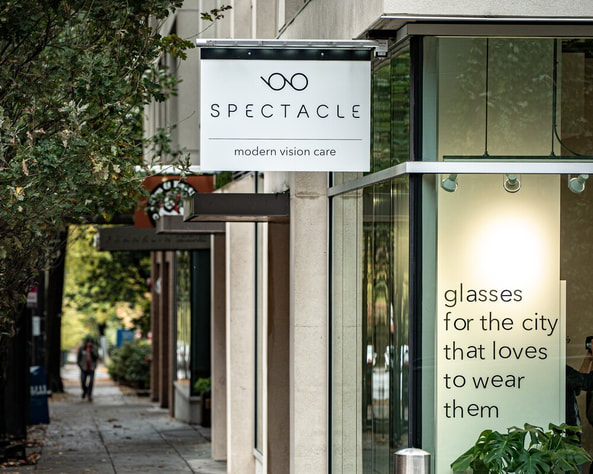 White Spectacle optometry sign with glasses icon on a modern city storefront.