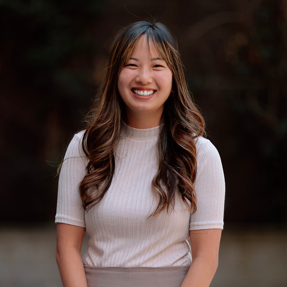 Our Team 5 Young Asian woman with long wavy brown hair, smiling broadly in a light top.