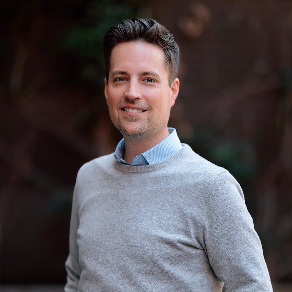 Our Team 1 Smiling man with dark hair and blue eyes wearing a grey sweater and blue shirt.