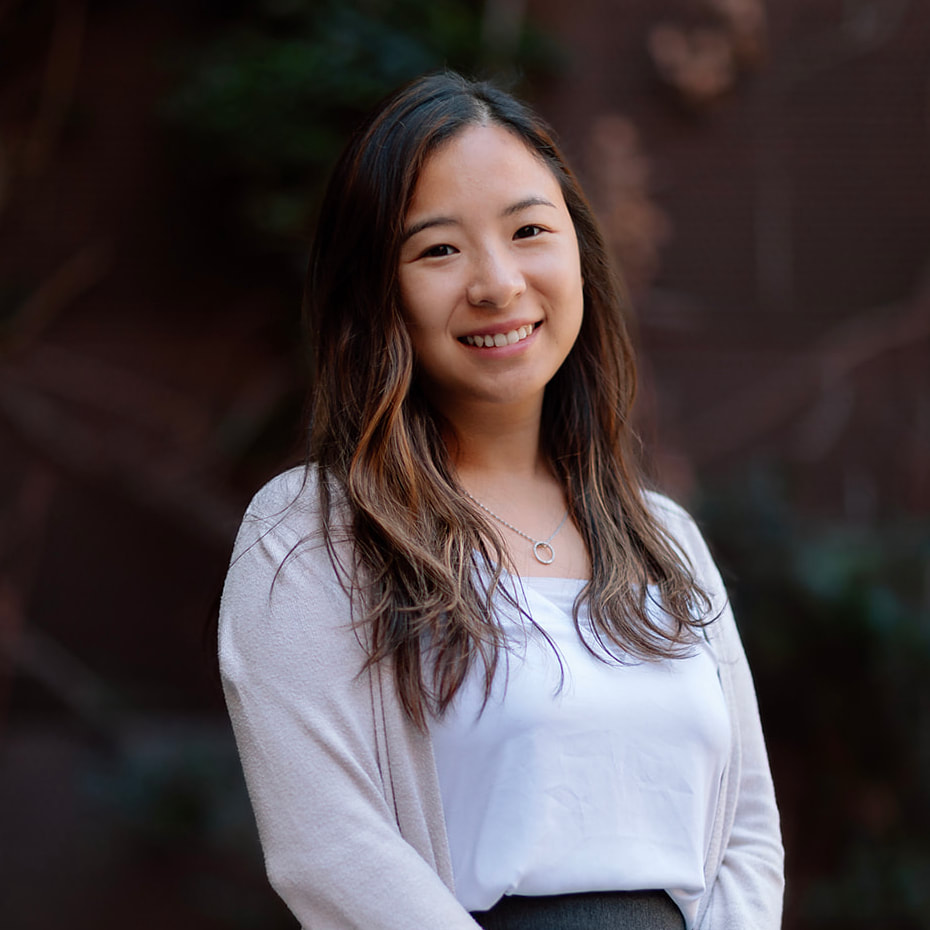 Our Team 2 Smiling young Asian woman with long wavy brown hair wearing a white top and cardigan.