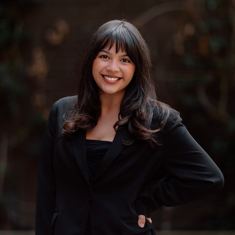 Our Team 11 Confident young woman with dark hair and bangs smiles warmly, wearing a black blazer.