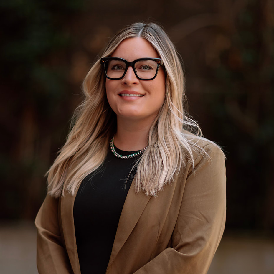 Our Team 6 Smiling blonde woman wearing black glasses, a silver necklace, and a brown blazer.