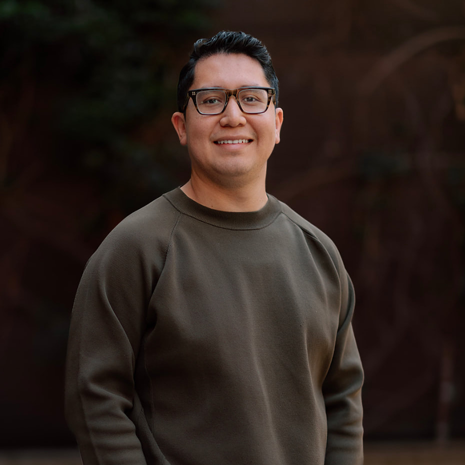 Our Team 10 Smiling man with dark hair, glasses, and a dark green sweater. Blurred background.