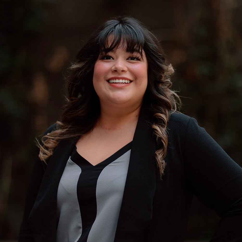Our Team 16 Smiling woman with dark, wavy hair and bangs, wearing a black blazer.