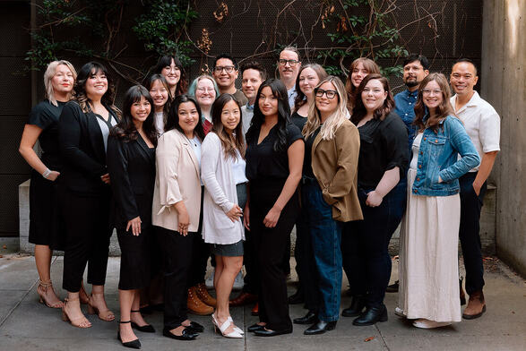 A smiling, diverse group of twenty adults stands outdoors in front of an ivy wall.