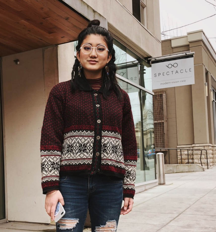 Smiling woman in round glasses, red snowflake cardigan, ripped jeans, outside an optician.