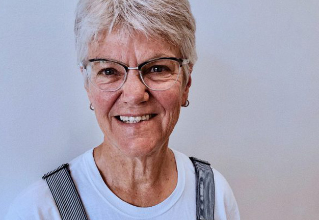 Home 13 Smiling older woman with short grey hair and glasses, wearing a white tee and striped overalls.